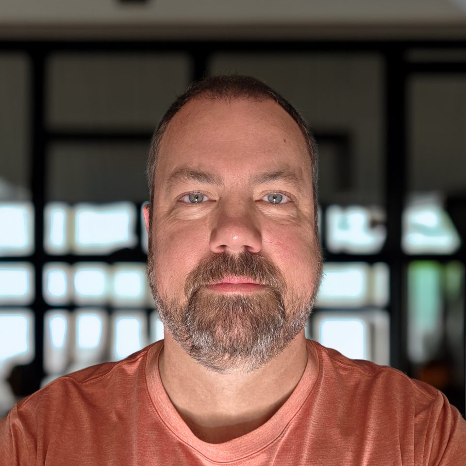Mike Ramsey Jr — a bearded man in a rust-coloured T-shirt, looking straight at the camera, in front of a softly blurred window.
